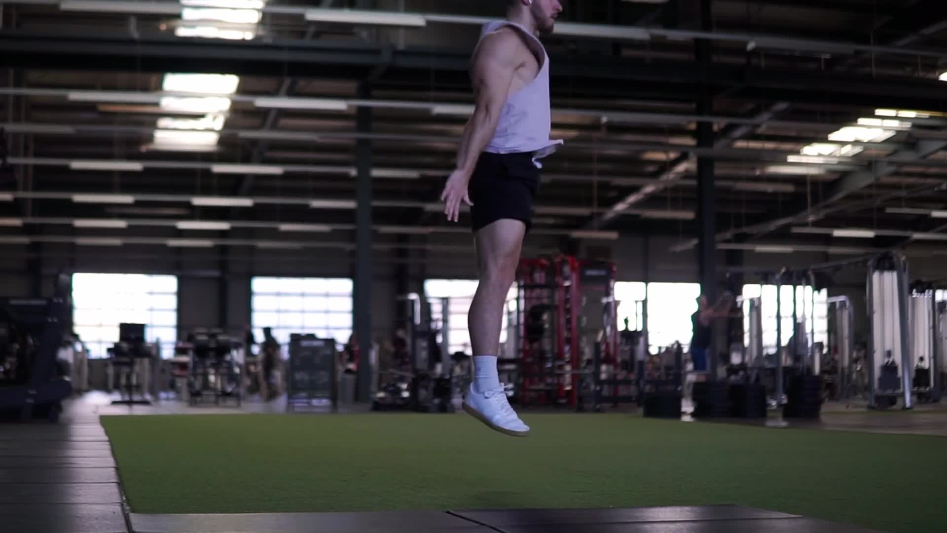 Male athlete performing an explosive squat jump in a minimalist gym with concrete wall and black rubber floor mats
